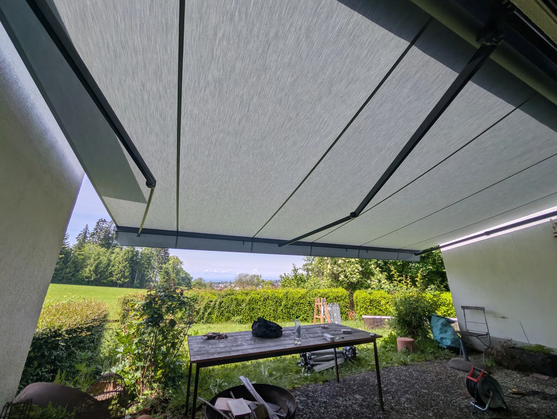 Große Gelenkarmmarkise über einer Terrasse, darunter ein Tisch, dahinter Wiese, Hecke und Blick ins Tal