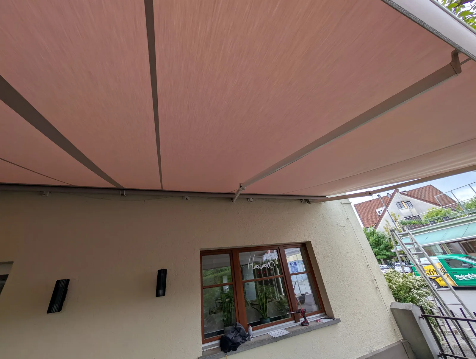 Zwei terrakottafarbene Markisen über dem Gastgarten eines Lokals an einer Straße