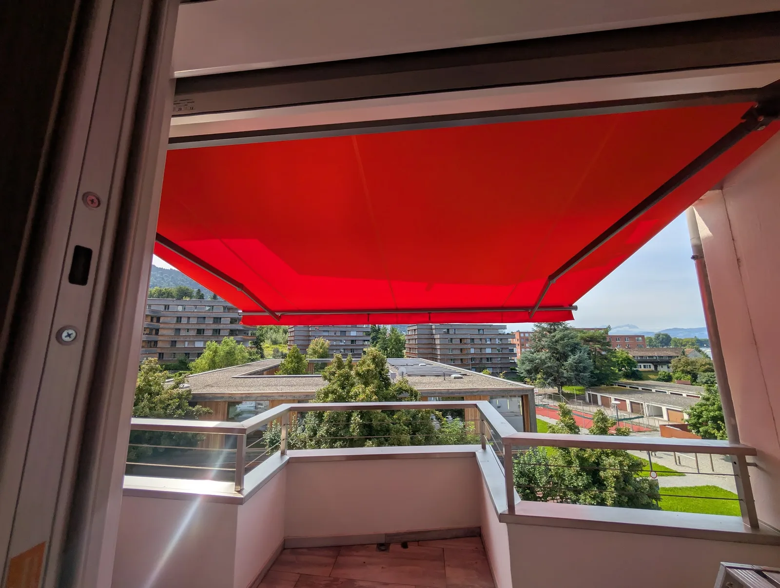 Rote Gelenkarmmarkise über einem Balkon, dahinter Wohnhäuser und Berge